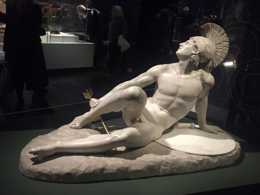 Status of Achilles, who is sitting naked on the ground as he grips the top of a long arrow that has pierced his right heel.
