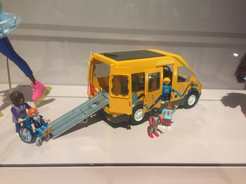 Small toy model of a yellow minibus, with a boy in a wheelchair about to be pushed up a ramp into the back of the bus by a woman. A few other characters are gathered together at the side of the bus, one of whom is waving.