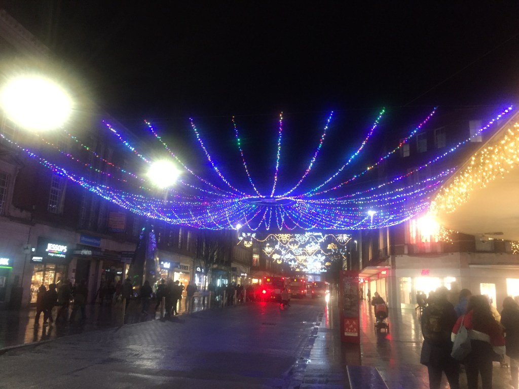 A huge chandelier-like structure, made of many lines of lights curving out and upwards from a central ring of light, spans the entire width of Exeter High Street. Other decorative Christmas lights can be seen across the street in the distance.