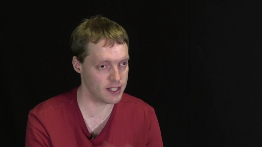 Glen wearing a red top while seated in front of a red background,