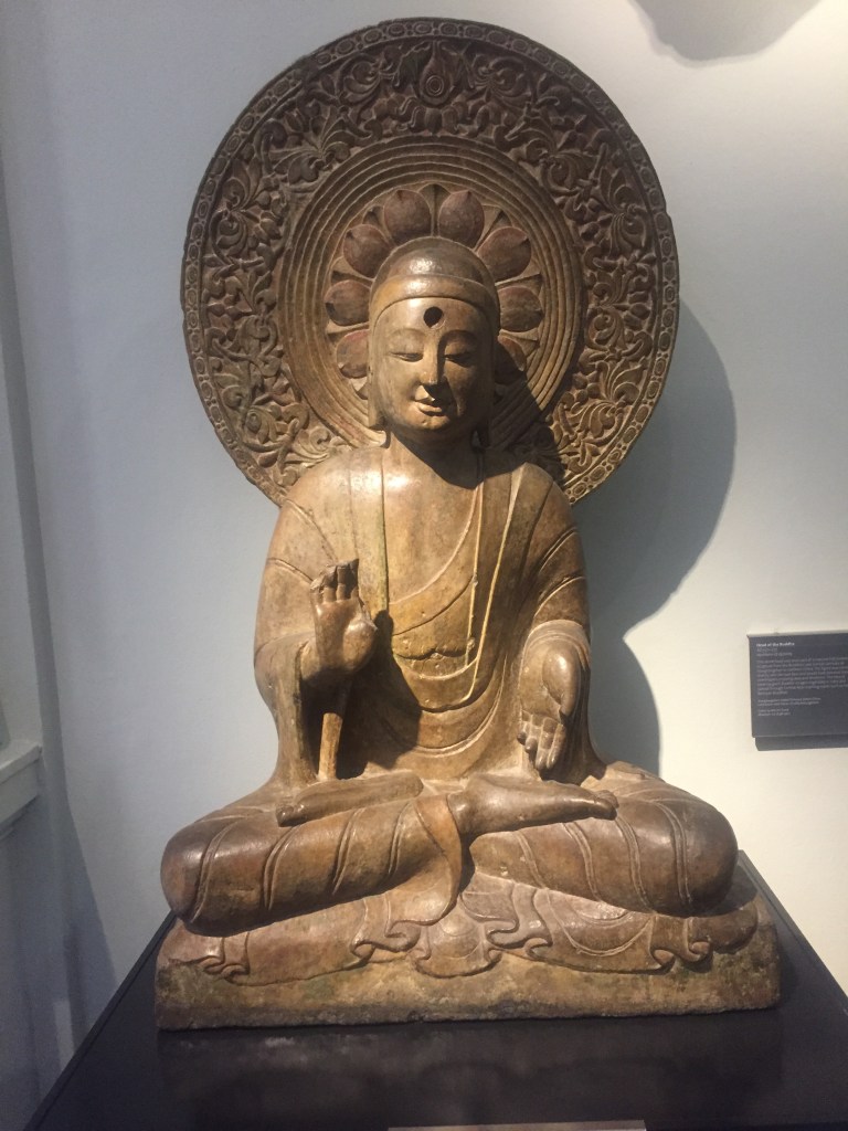 Statue of the Buddha seated in a meditation position, with their legs crossed so that their knees are pointing out to the sides. Behind their head is a large and ornately decorated circular shield. The leaf-like shapes around the central section give the impression that the Buddha's head is in the middle of a flower.