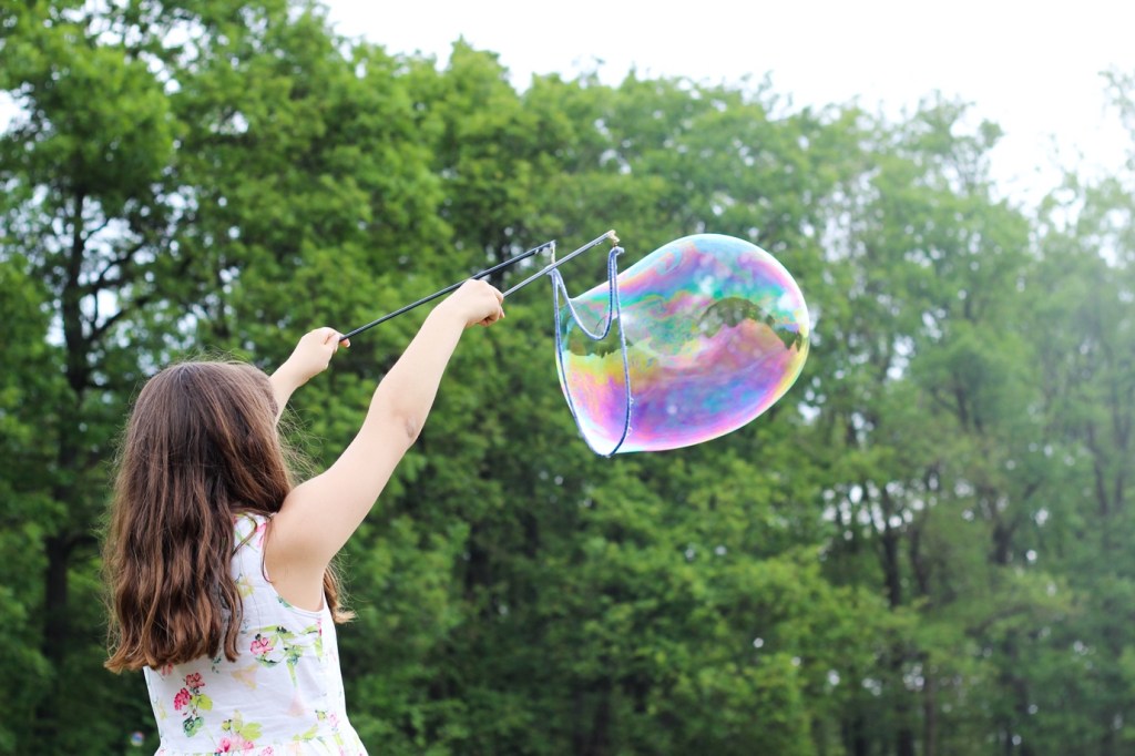 A young girl outdoors using a giant bubble wand, producing a large bubble that has a rainbow-coloured effect as it reflects the light.