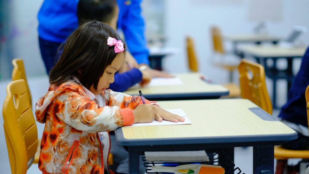 A young girl, wearing a jacket covered in orange flowers and a pink bow in her hair, drawing on a piece of paper while sat at a school desk.