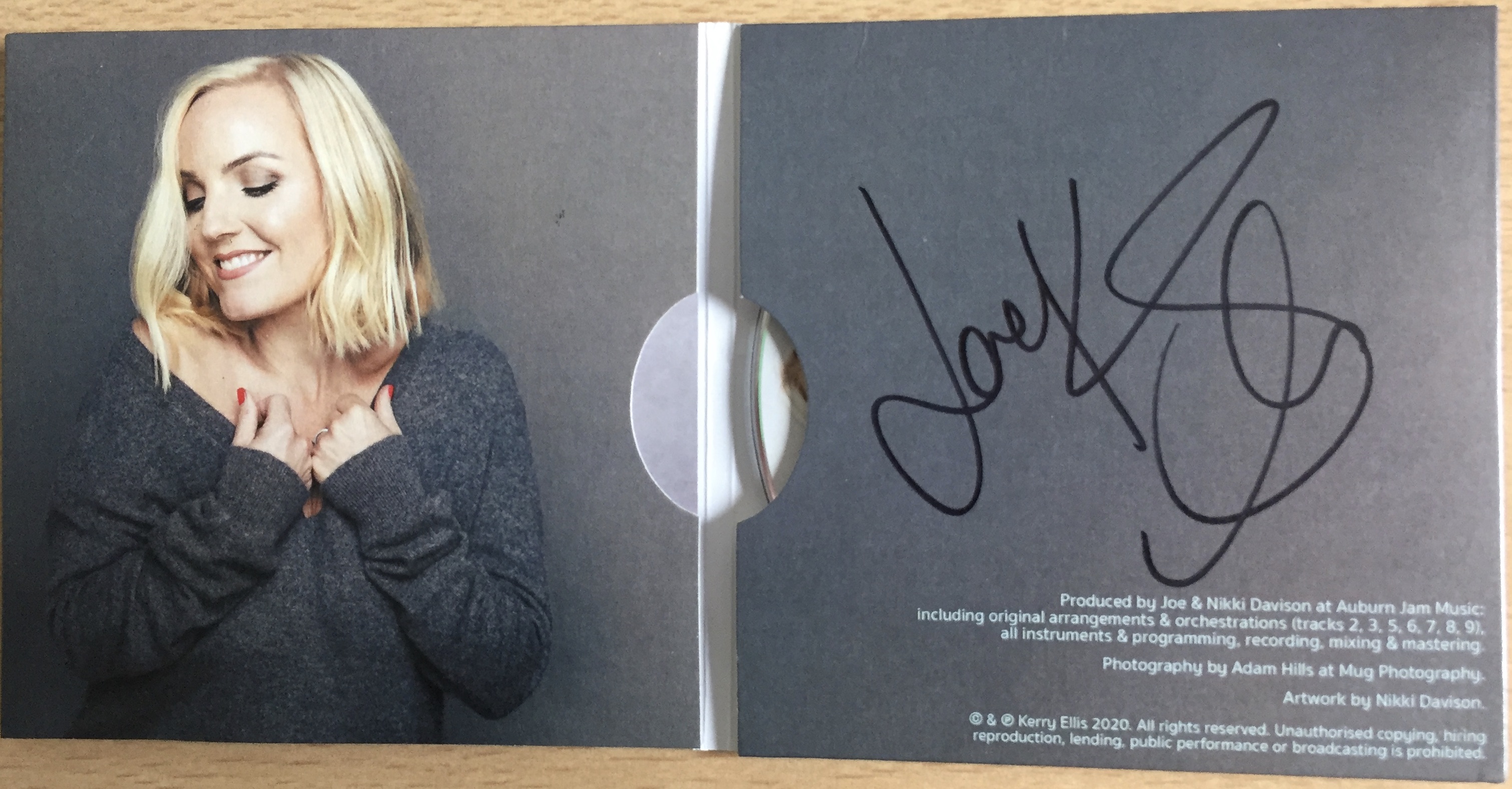 The unfolded sleeve for the Kerry Ellis album Feels Like Home. On the left side is the blonde singer smiling with her eye closed and her hands holding the top of her grey jumper. On a grey background on the right side is Kerry's signature in black, filling three quarters of the space, above small credits in white text in the bottom right corner.