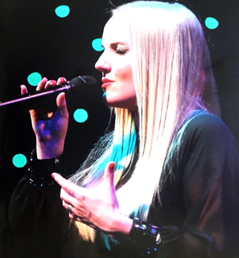 Kerry Ellis singing live on stage. She has long blonde hair, red fingernails and is wearing a black top.
