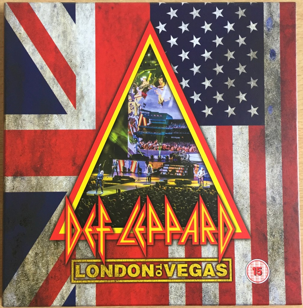 Front cover of the Def Leppard London To Vegas box set. The background is split into 2 vertical sections, containing half of the Union Jack and American flags respectively. A large triangle in the middle, with a double border of red and yellow, features a colourful photo of the band in concert, with singer Joe Elliot on a platform stretching out from the stage into the audience. Across the bottom of the triangle, large yellow text with a red border says Def Leppard, in a font made only of straight and diagonal lines. Below this, in a yellow box, yellow text says London To Vegas.