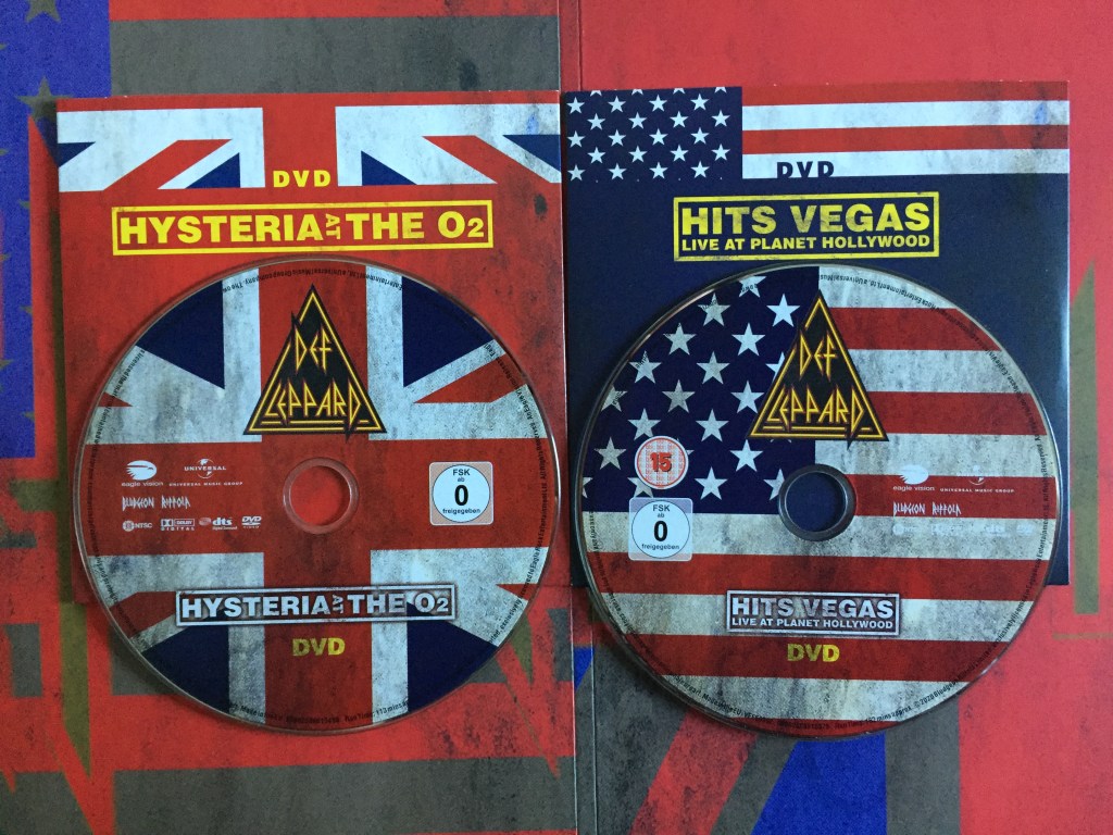 The DVD discs for Hysteria At The O2 and Hits Vegas Live At Planet Hollywood by Def Leppard. The background of each disc shows the central portion of the Union Jack or American flag respectively. The Def Leppard logo, made of a red and yellow bordered triangle with the yellow lettering of the band name filling the centre, sits in the top centre of the disc. The show name is in the bottom section of the disc, as yellow text inside a yellow bordered box.
