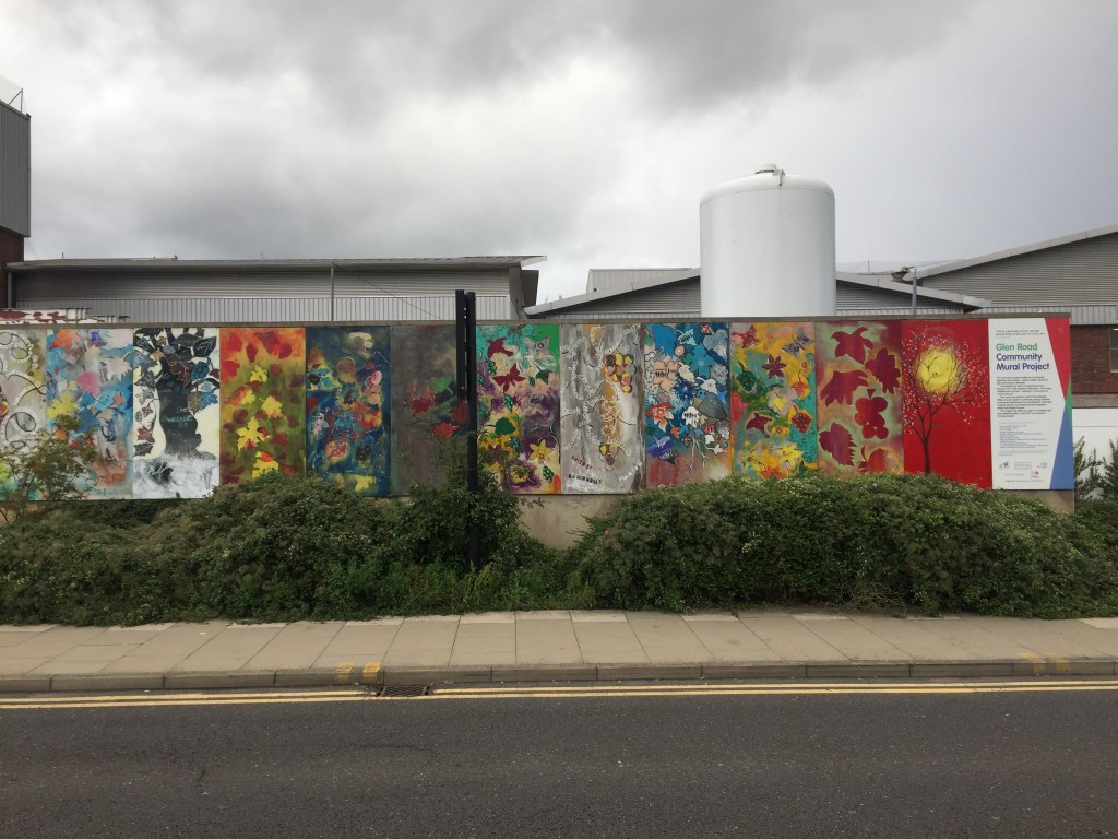 A long mural stretching along a wall by the road, made of several vertical panels, each containing a different piece of colourfully painted artwork. They all have a nature theme, with leaves, flowers, trees, birds, sunshine, etc. The final panel on the right shows that this is titled the Glen Road Community Mural Project.
