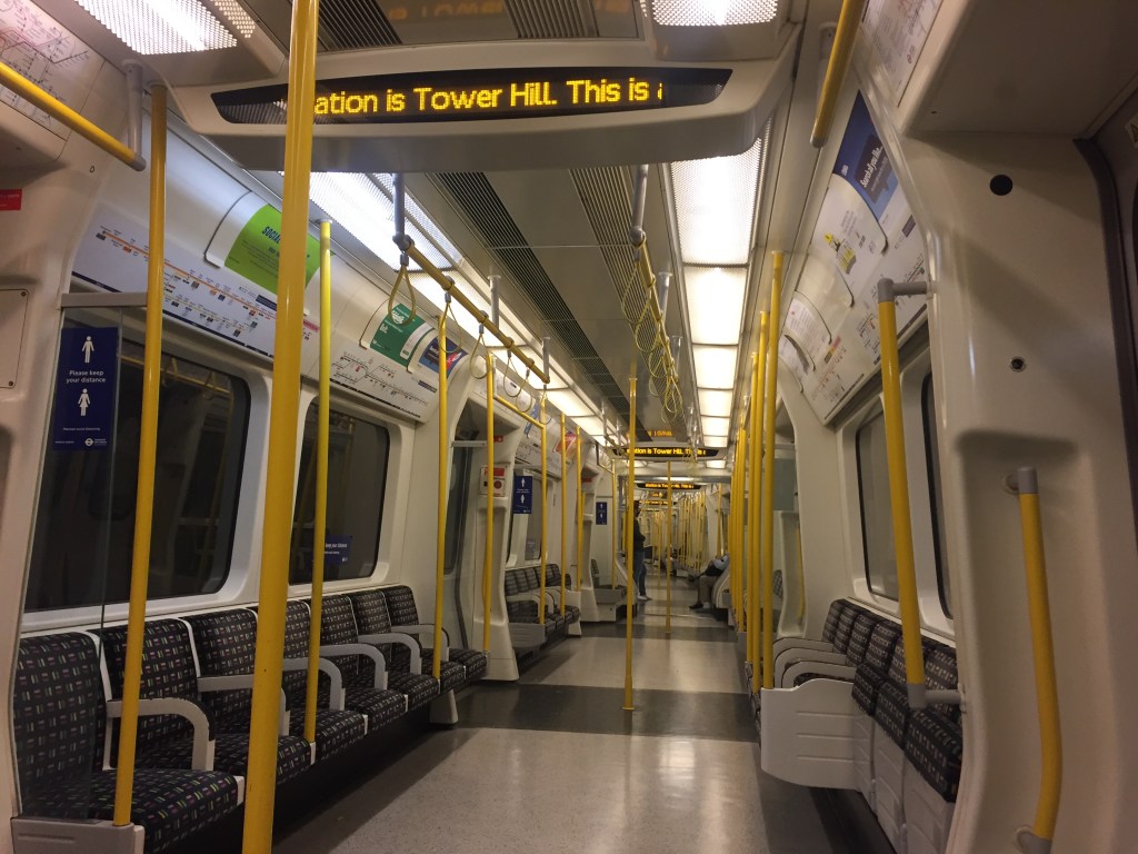 Inside a District Line train, where the entire section of the carriage ahead is empty, and only a few people are visible in the next section that's curving away in the distance. Dark blue signs on panels near the doors feature white male and female figures with the text Please keep your distance.