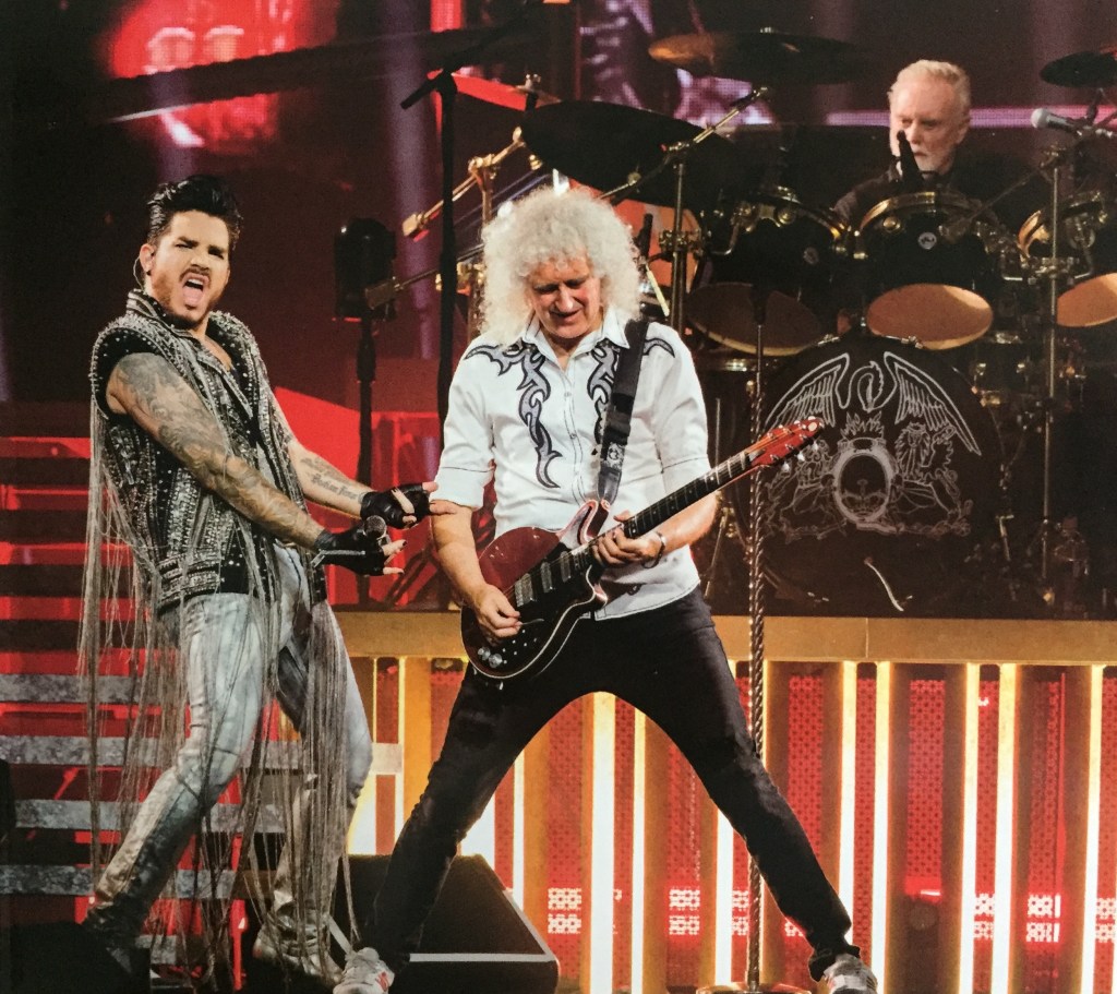 Brian May smiling and standing with legs wide apart as he plays the guitar. Adam Lambert, wearing a top with lots of chains hanging down to the floor and the short sleeves revealing his tattooed arms, is making a thrusting motion towards Brian, while shouting something towards the audience. Roger Taylor is on the drums in the background.