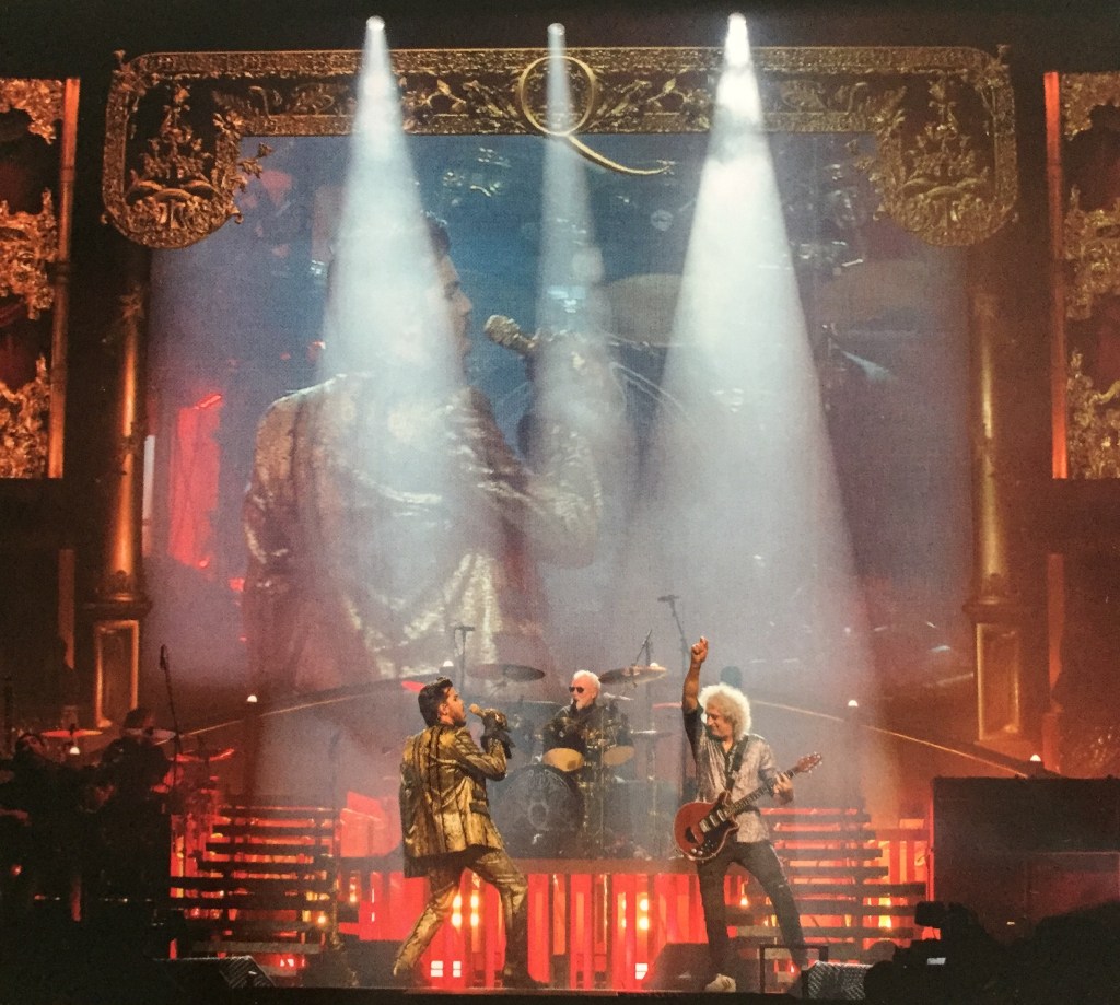 Queen & Adam Lambert performing on stage. Spotlights are shining down on Adam, Brian and Roger, while the ornately gold framed big screen at the back of the stage, with a big letter Q at the top centre of the frame, shows a close-up of Adam singing.