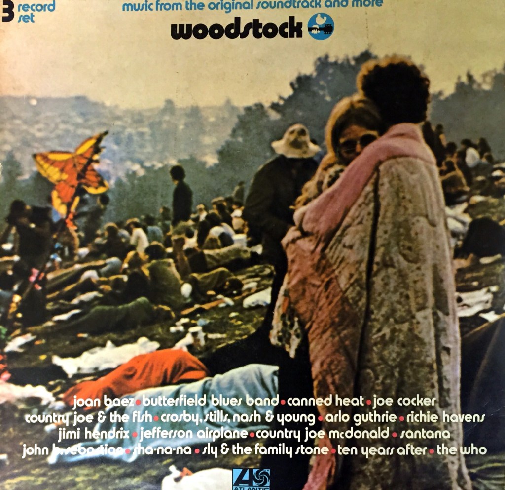 Woodstuck album cover, showing 2 people standing and embracing, while many others are laying down on the grass in the sunshine behind them.