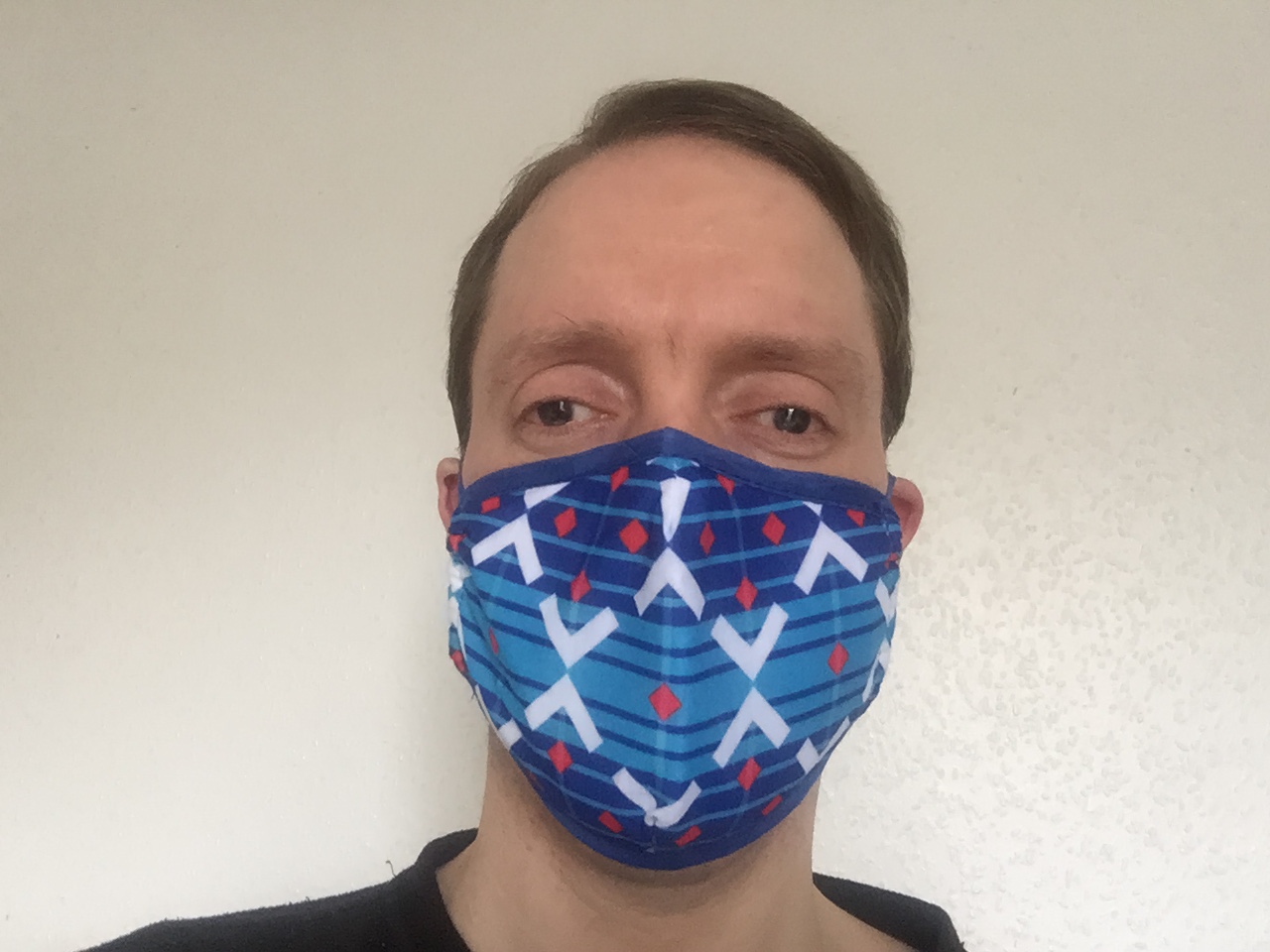 Selfie of Glen wearing a face mask decorated with a Victoria Line train moquette pattern. The pattern has alternating light blue and dark blue horizontal stripes of varying widths, overlaid with two zig-zagging white lines that form 3 diamond shapes, and there are small red diamonds spread around the pattern as well.