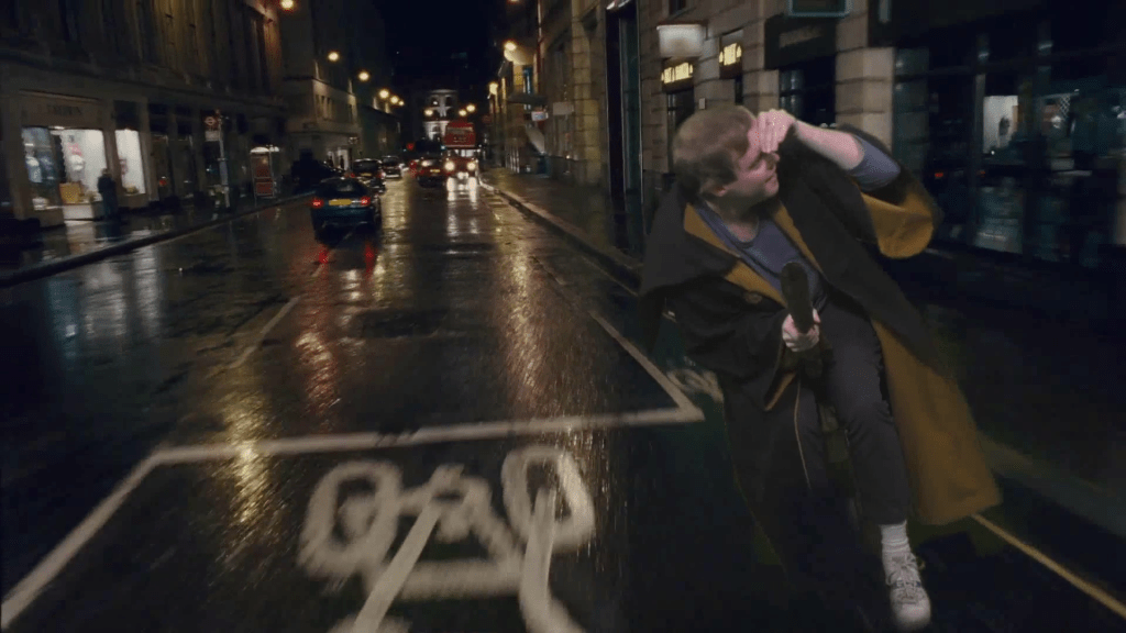 Glen looking around as he flies down a busy London street at night on a broomstick.