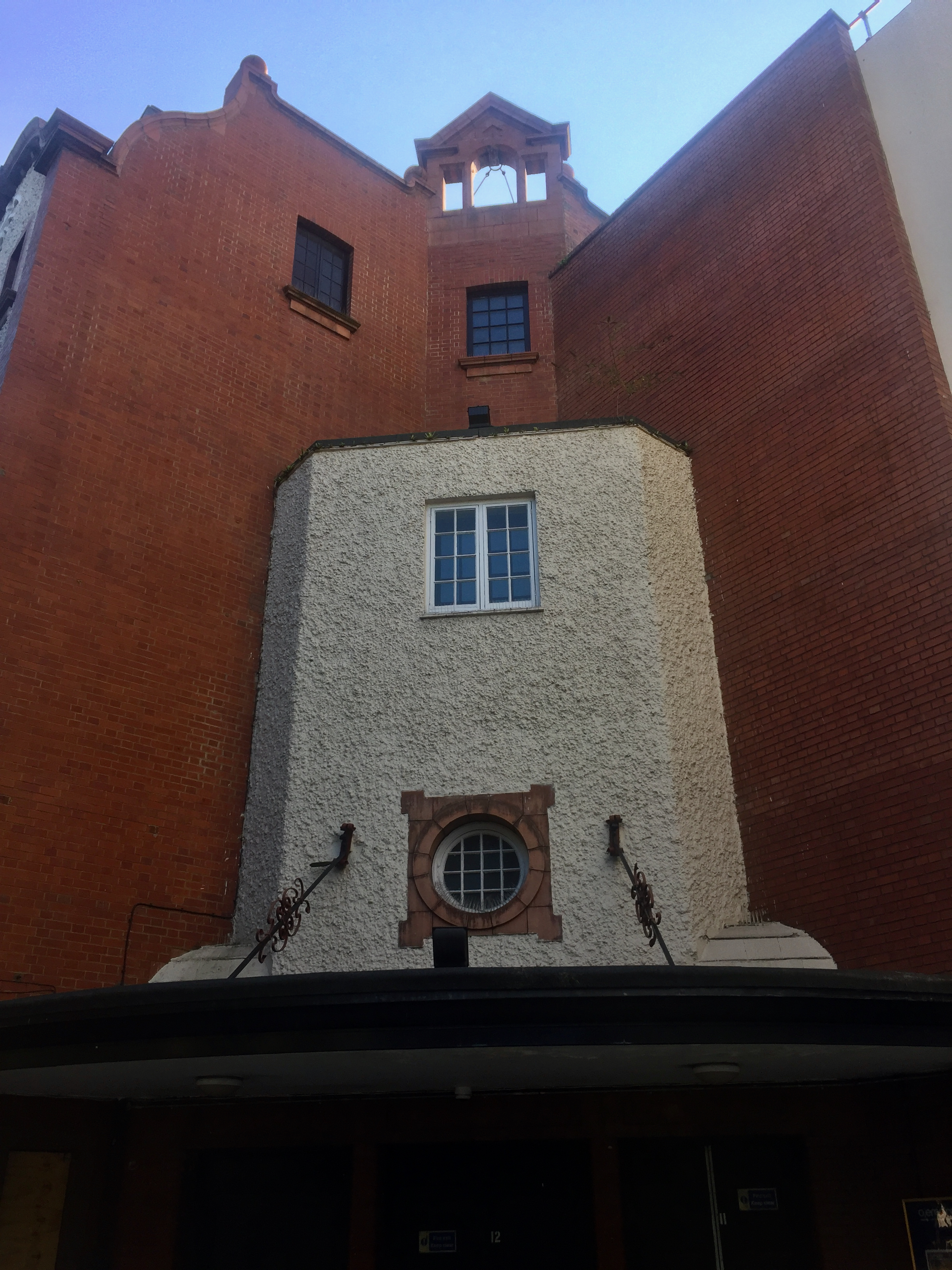 The front right corner of the Shepherd's Bush Empire. Above the ground floor, a white stone section has a small circular window on the first floor, and a normal vertical window on the second. This stone section is surrounded to the sides and one floor above by the red bricks that make up most of the venue's appearance.