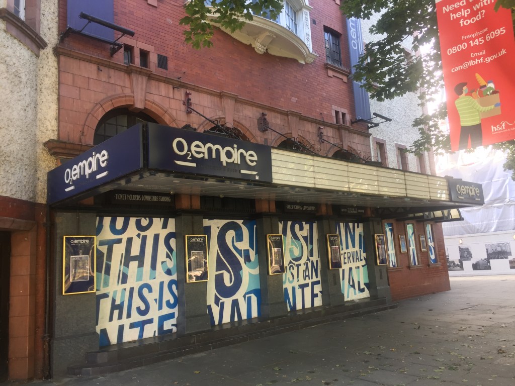 The front of the Shepherd's Bush Empire, now called the O2 Empire. White posters cover up the 4 windows along the front, with stylistic blue lettering that reads This Is Just An Interval, in reference to the shutdown enforced by the Covid pandemic.