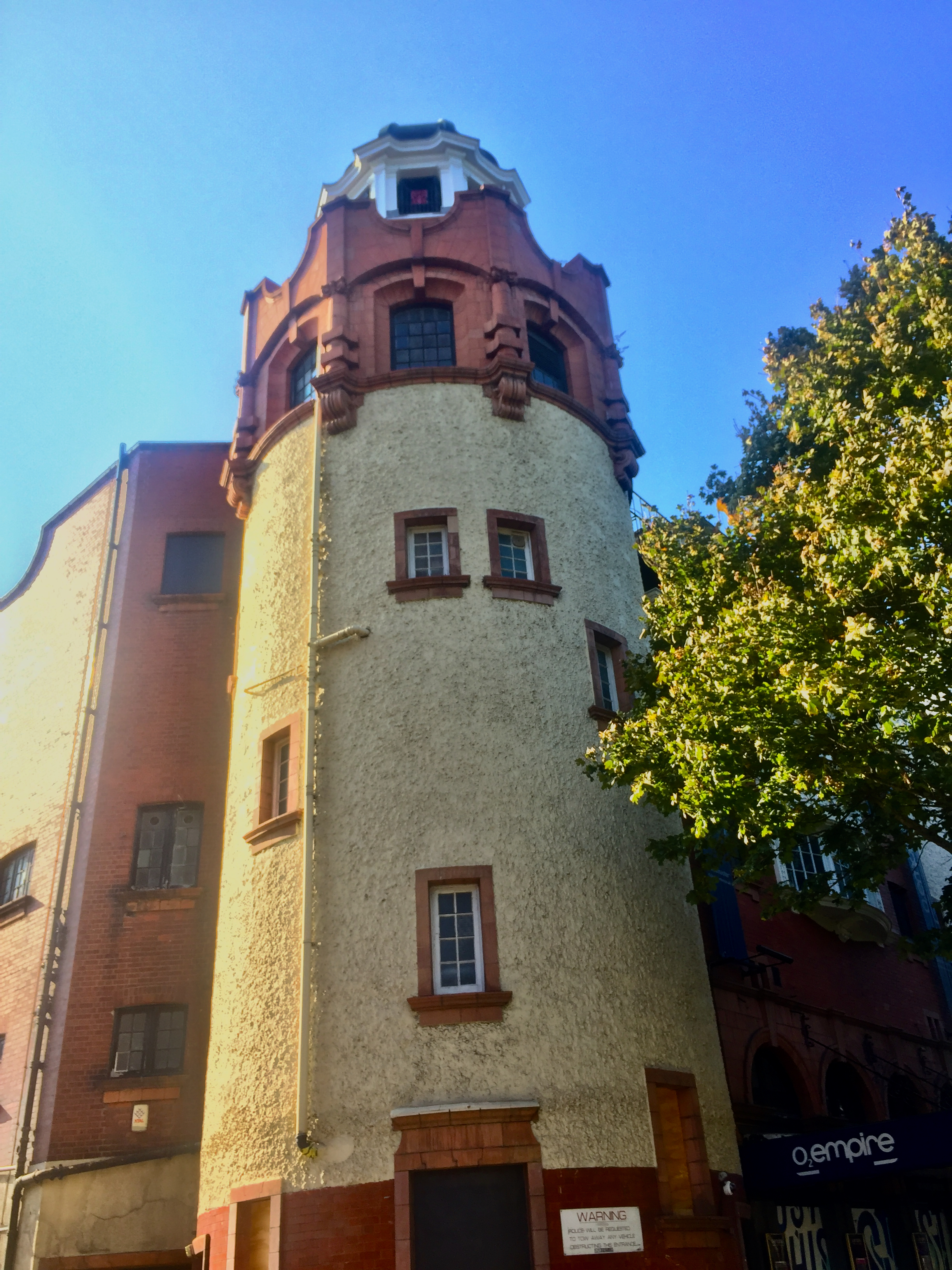 Tower on the front left corner of the Shepherd's Bush Empire. Above the ground floor, a white stone section forms the first 3 floors, with a few vertical windows in somewhat random positions around it. This is topped by a red brick section, with vertical windows all around the edge at regular intervals, and at the very top is a smaller white domed section, again with windows all around it.