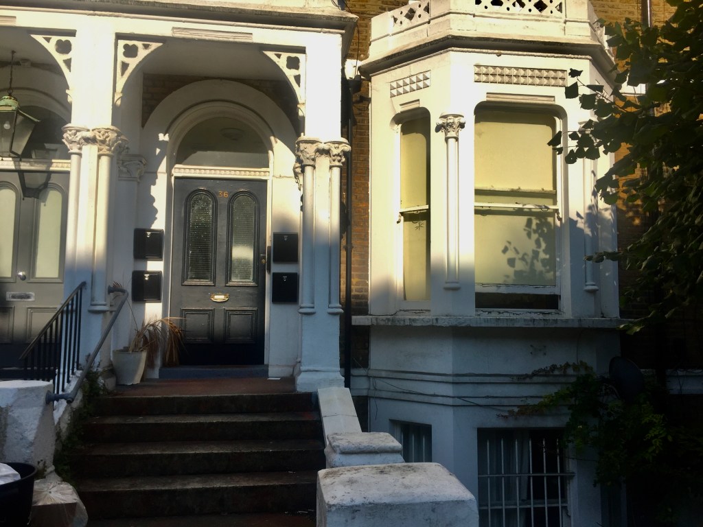 The ground floor of number 36 Sinclair Road, with white framing around the porch, door and windows. The front door is black or dark grey, with 2 tall arched and frosted windows on its top half.