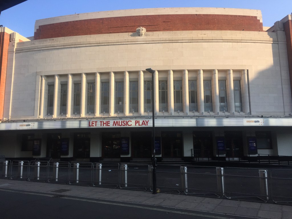 The Eventim Apollo, a large, wide building. Above the ground floor, which has steps leading up to multiple entrance doors, a narrow panel across the entire building features the venue's name and large red letters reading Let The Music Play. Above that, the building is mainly large white concrete, with a row of 14 tall, narrow windows stretching about halfway up. The very top of the venue has a red brick layer topped by a small layer of white concrete.