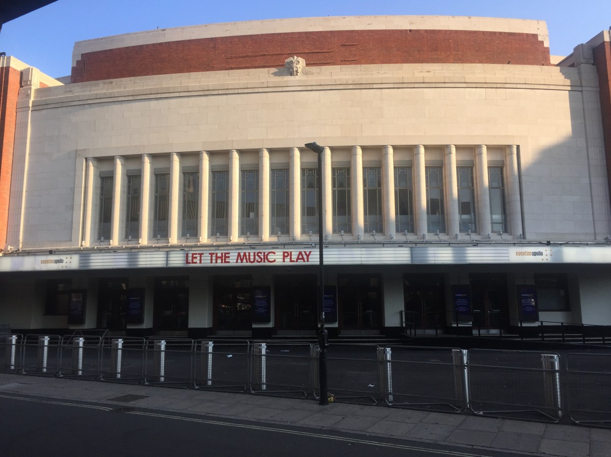 The Eventim Apollo, a large, wide building. Above the ground floor, which has steps leading up to multiple entrance doors, a narrow panel across the entire building features the venue's name and large red letters reading Let The Music Play. Above that, the building is mainly large white concrete, with a row of 14 tall, narrow windows stretching about halfway up. The very top of the venue has a red brick layer topped by a small layer of white concrete.