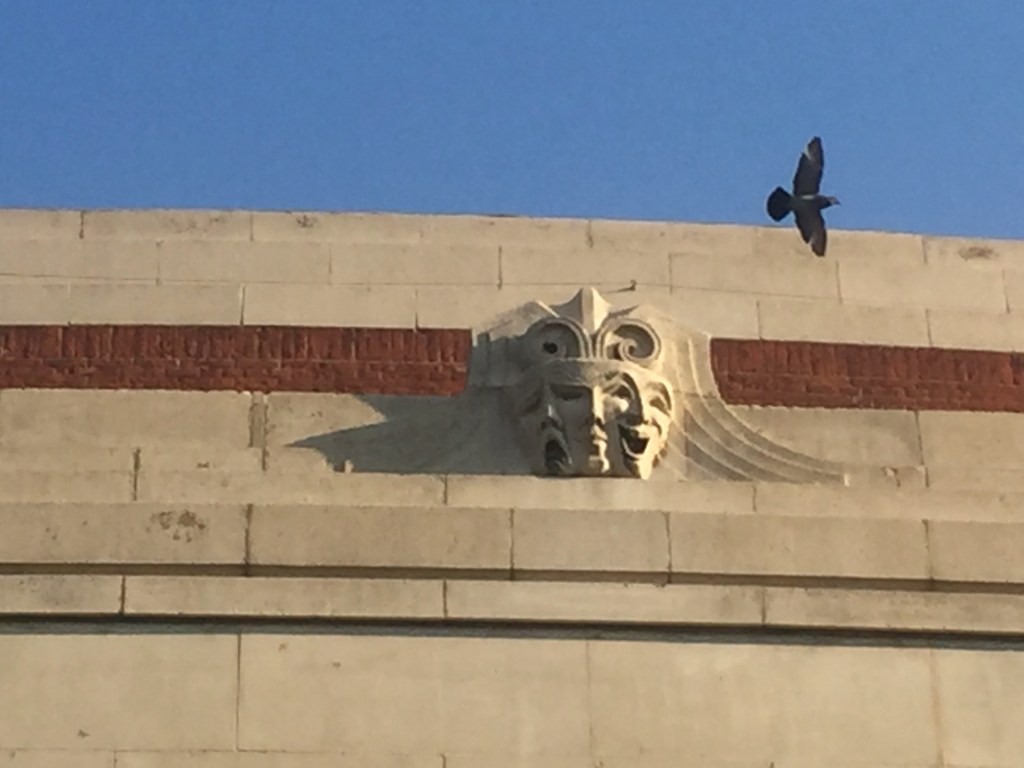 Close up of 3 theatrical faces sculpted into the concrete at the top of the Eventim Apollo, one with an exaggerated expression of sadness, one looking unemotional, and one looking very happy. A black coloured bird is flying past, its wings outstretched.