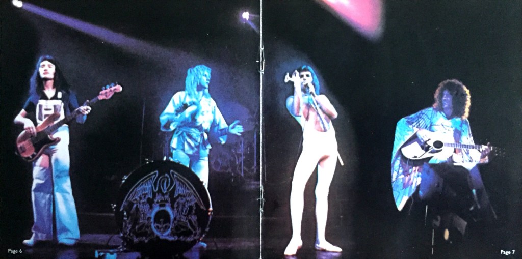 Centre spread from the booklet in A Day At The Races, showing a photo of Queen performing on stage. From left to right, John is playing bass guitar, Roger is standing by a small bass drum while playing a tambourine, Freddie is singing in his white low-cut jumpsuit, and Brian is sitting on a stool playing acoustic guitar.