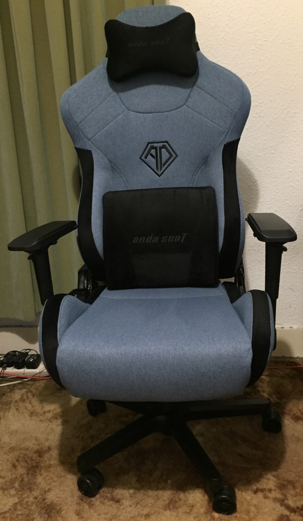 The Andaseat T-Pro 2 gaming chair, a blue chair with a high back, adjustable black armrests, a black lumbar pillow for the lower back and a smaller black headrest pillow strapped around the top of the chair.