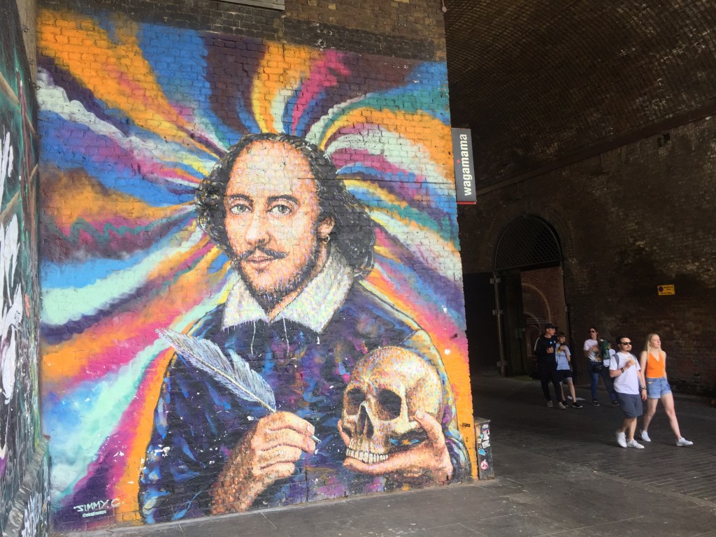 A large mural of William Shakespeare holding a quill in his right hand and a skull in his left, against a colourful swirling background.