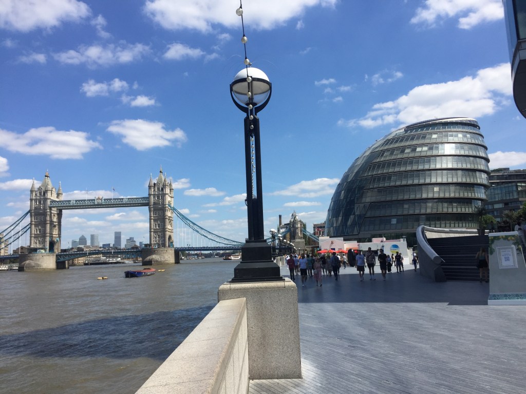 View along the South Bank, showing Tower Bridge on the left, a lamppost on the wall in the centre, people walking the path to the right, and the rounded structure of City Hall on the far right.