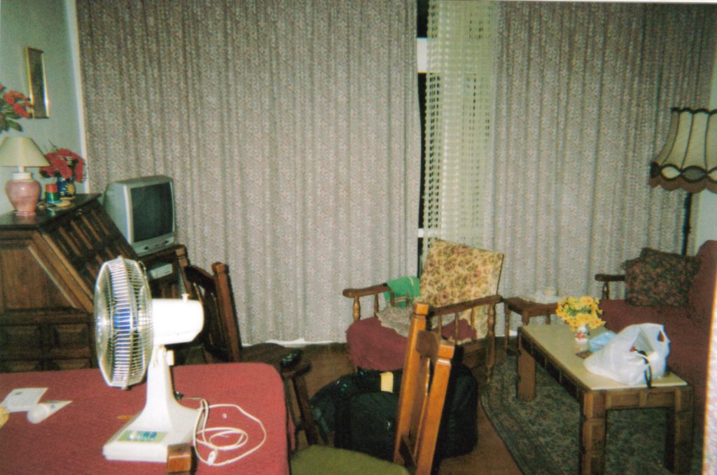 The lounge in my Spanish apartment, including tables and chairs, a portable electric fan, a TV in the corner, and curtains across the doors to the balcony.