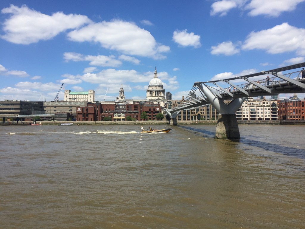 A speedboat containing 4 people passing under the Millennium Bridge, with St Paul's Cathedral in the background.