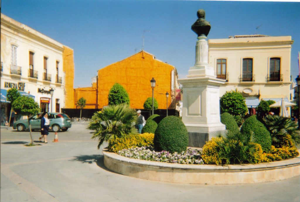 A circular flowerbed in the town in Ronda with greenery and other plants. In the centre is a tall white pedestal with a bronze bust of someone's head and shoulders on the top.