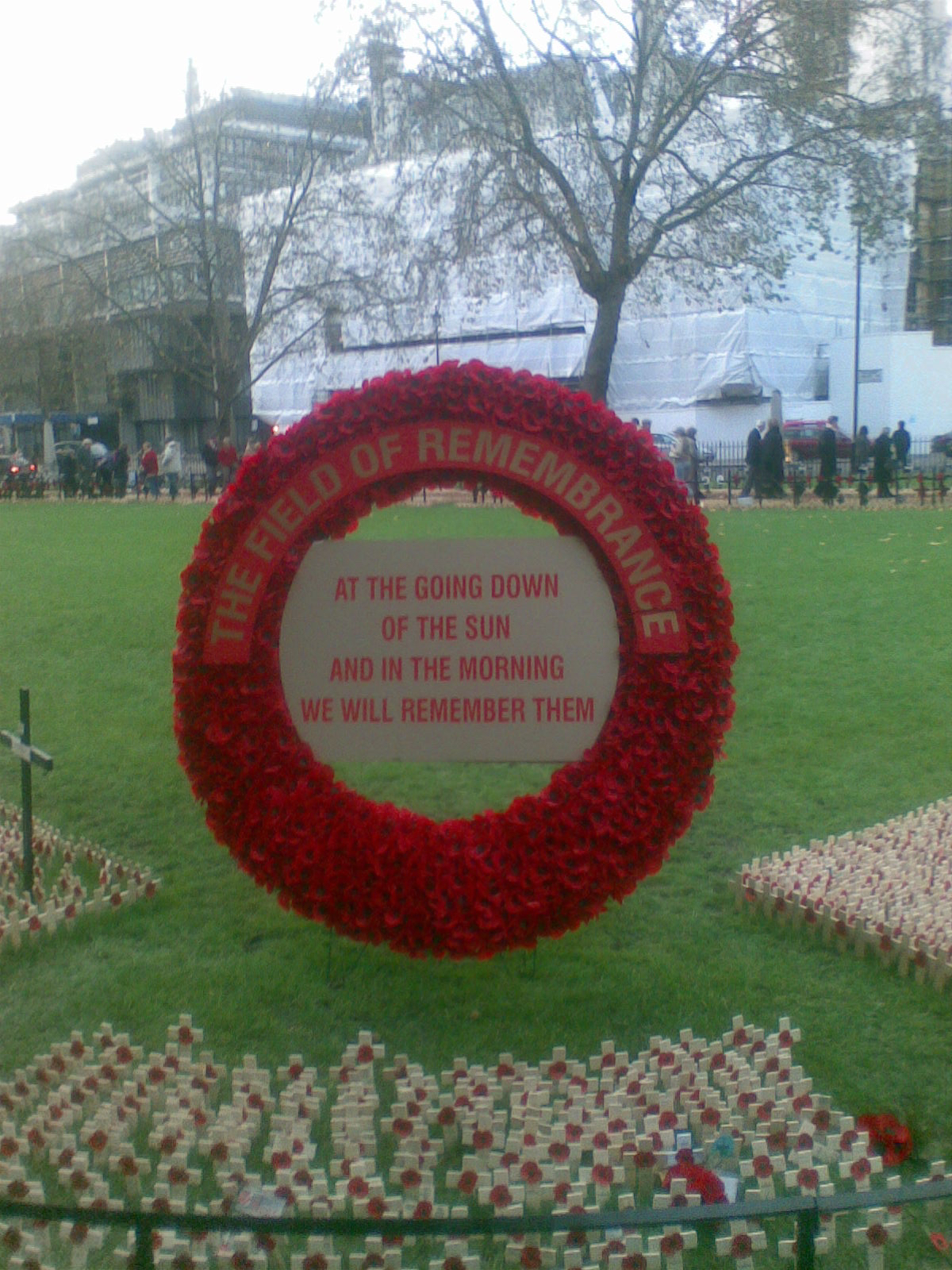 Large poppy wreath sign in front of a large grassy area. Text arching around the top reads The Field of Remembrance. A plaque in the centre of the wreath reads at the going down of the sun, and in the morning, we will remember them.