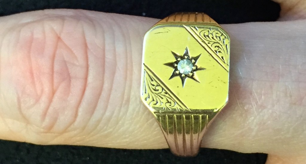 Gold signet ring, in a square shape with rounded corners, with the top right and bottom left corners having an ornate wavy design. An 8-point star in the centre of the ring holds a small diamond.