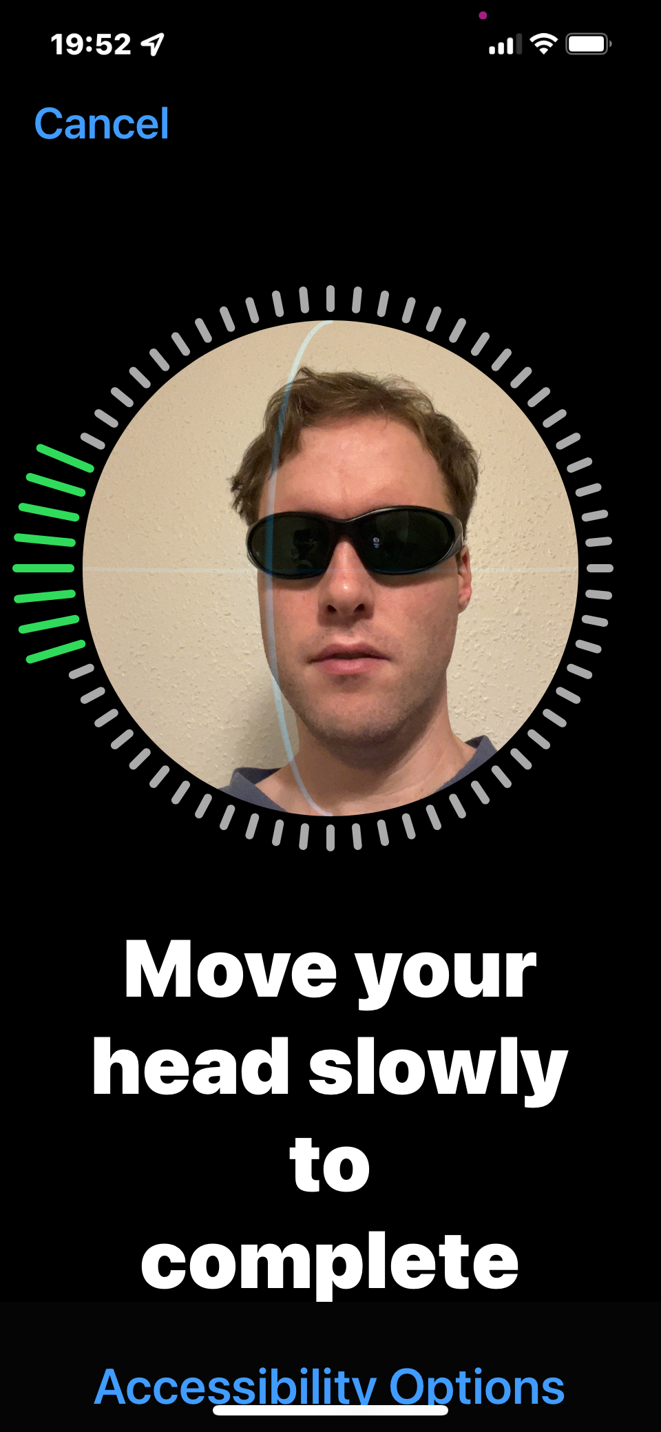 iPhone 13 screenshot. My face with sunglasses on is positioned inside a circular frame. All around the frame are small grey bars, which turn into longer green bars as I turn my head to help the phone recognise me. Large text below the frame says Move your head slowly to complete, along with a link for accessibility options.