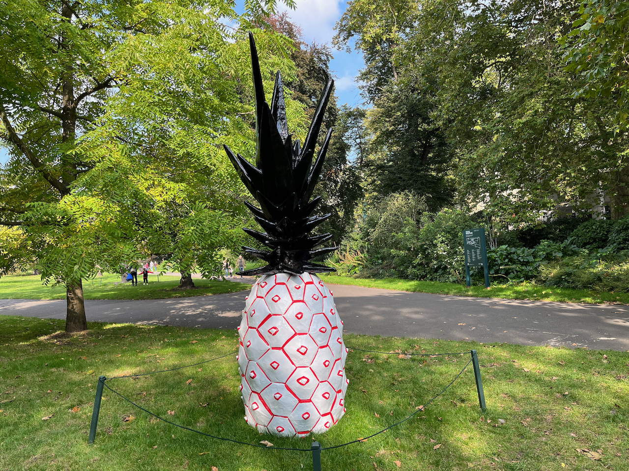 Large sculpture of a pineapple in Regent's park. The body is white with a pattern of red hexagonal outlines, while there are tall thick black leaves sticking out of the top.