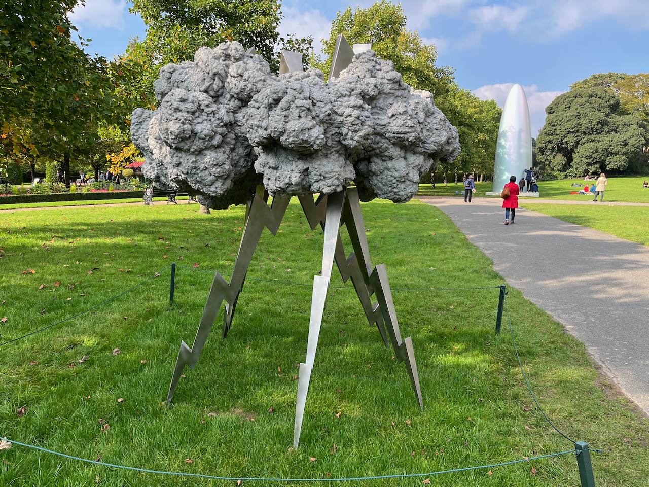 Sculpture of a big grey thunder cloud, stood on 4 legs of thick zig-zag lightning on the grass. In the background, at the end of the path, the sun is shining on a huge metal bullet-shaped sculpture.