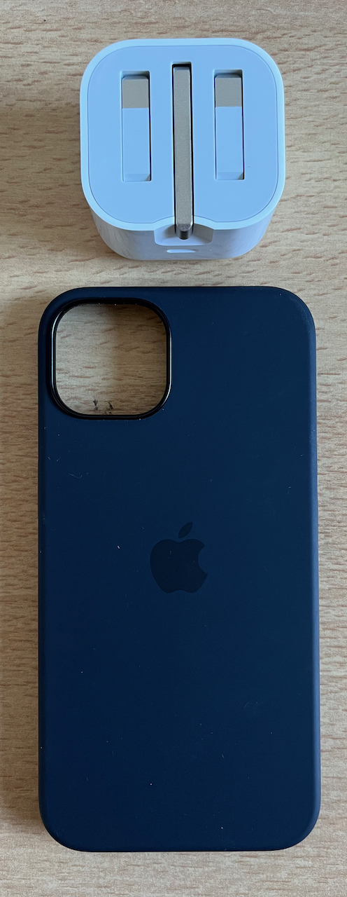 iPhone 13 charger plug in white, with the 3 metal prongs folded inwards to keep it flat during transportation. And a black silicone phone case, with an Apple logo in the centre and a hole in the top left for the cameras.