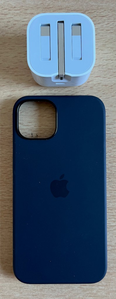 iPhone 13 charger plug in white, with the 3 metal prongs folded inwards to keep it flat during transportation. And a black silicone phone case, with an Apple logo in the centre and a hole in the top left for the cameras.