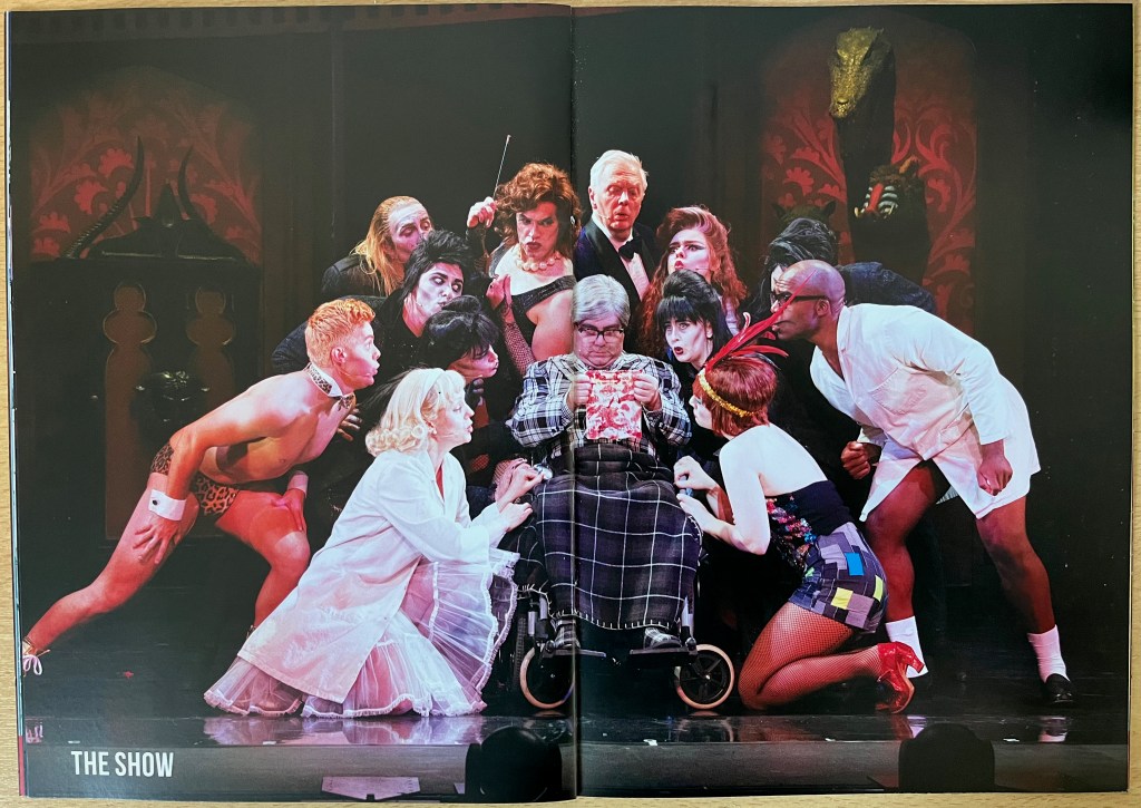 Double-page spread from the Rocky Horror tour brochure, with a single large photo showing the cast crowding around Doctor Everett Scott as he sits in his wheelchair and reads a letter.