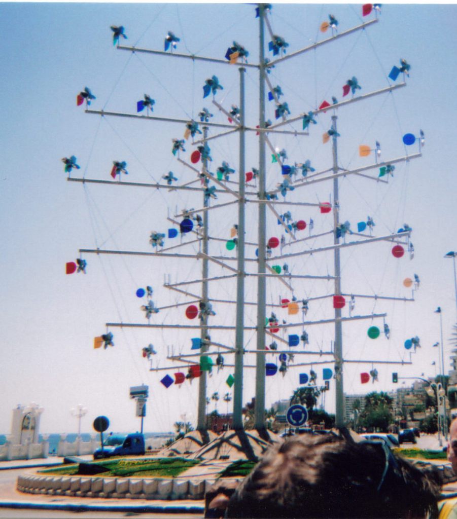 A tall metal structure in the middle of a small roundabout. Protruding from top to bottom of 4 tall poles are lots of long, thin horizontal rails, each of which has small colourful windmills attached.