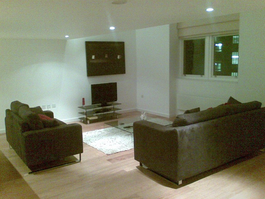 Spacious lounge area in a white-walled apartment, with spot lighting in the ceiling. A low, square glass table sits on a white rug in the centre of the space. Around it are a flat-screen TV on a low stand below a large framed picture on the wall, a 2-seater sofa, a 3-seater sofa, and 2 adjacent windows through which the lit windows of other apartments can be seen.