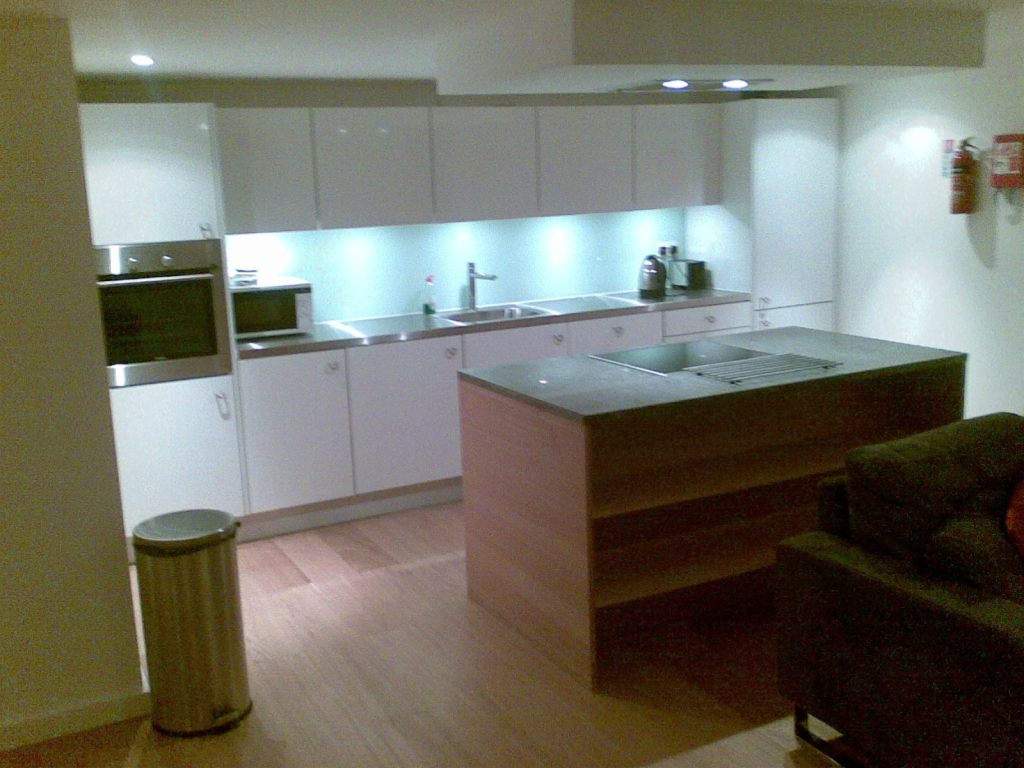 Kitchen area in a white-walled apartment, and spot lighting in the ceiling. The white kitchen units are along one wall. On the left, a big metal oven has a cupboard above and below it. To its right, a metal worktop has a microwave, sink, kettle and toaster. Below it are 5 cupboards and a set of drawers, while above are 6 cupboards. And on the far right is a tall fridge freezer. A large square wood-coloured unit, with 3 shelves on the side facing us, sits in the middle of the kitchen area, separate from the main units, with a hob visible on the top on the far side nearest the kitchen units.