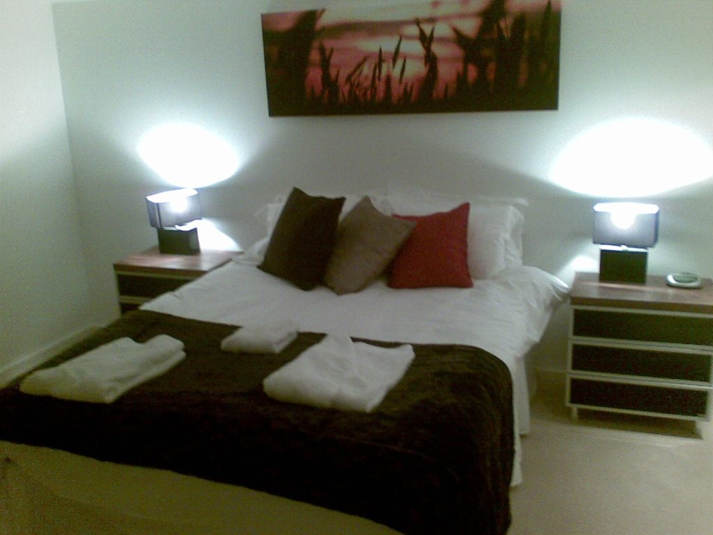 Bedroom in a white-walled apartment. The double-bed has white sheeting and pillows. 3 different coloured square cushions sit together in front of the pillows, while 3 different sized towels are folded and placed on a dark blanket covering the bottom half of the bed. A bedside table with 3 drawers sits on each side of the bed, each with a lamp on top with a square lampshade. On the wall above the bed is a large picture of grass or leafy plants in front of a sunset.