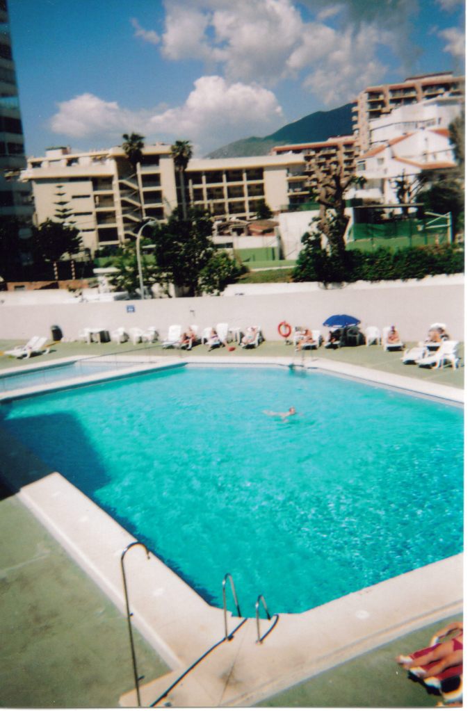 Outdoor swimming pool in a hotel complex, with people relaxing on sun loungers by a nearby wall in the sunshine.