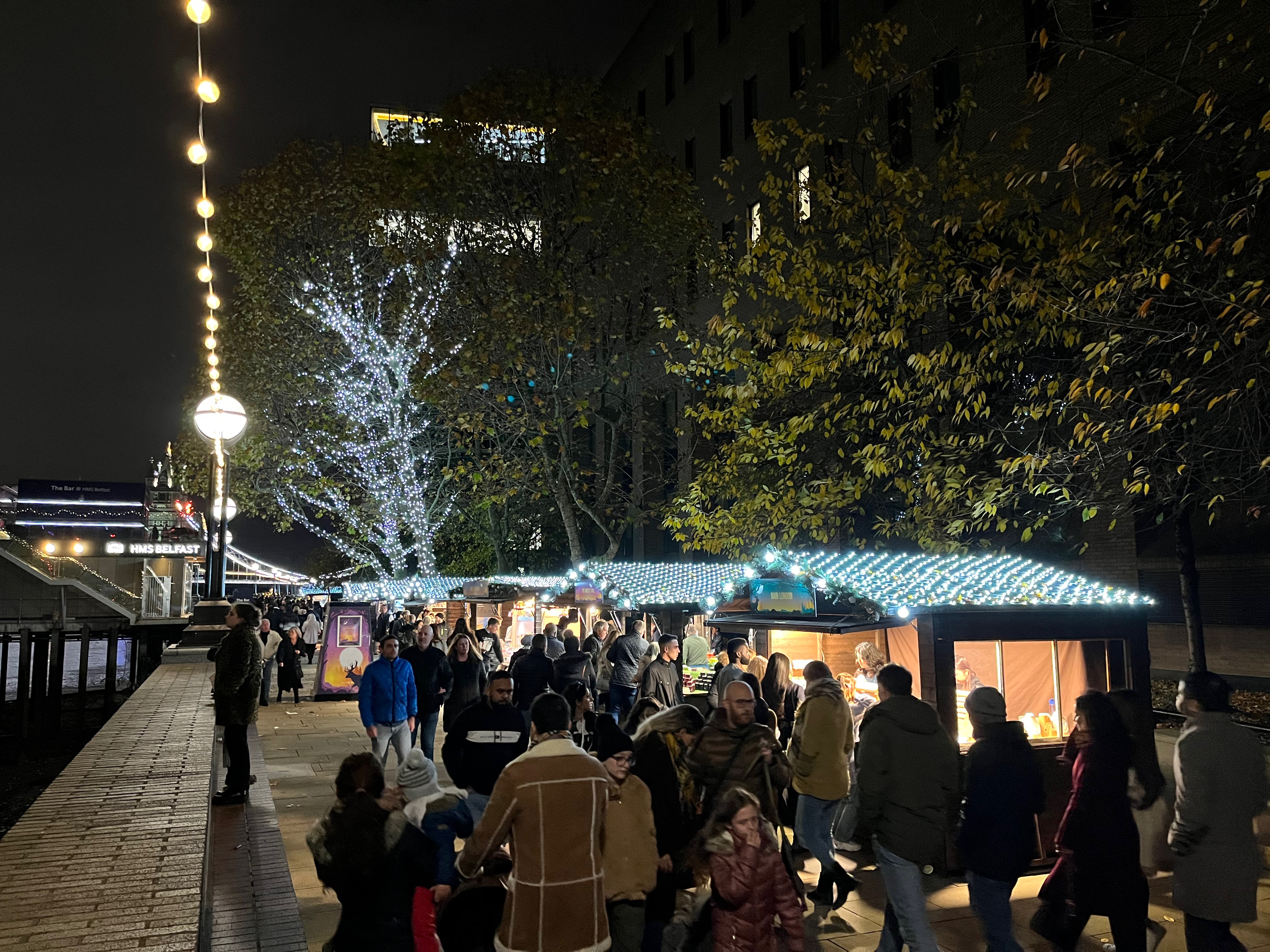 People shopping at street food stalls along the South Bank. The roofs of the stalls, along with a few tall trees in the distance, are covered in bright white lights to make them look festive.
