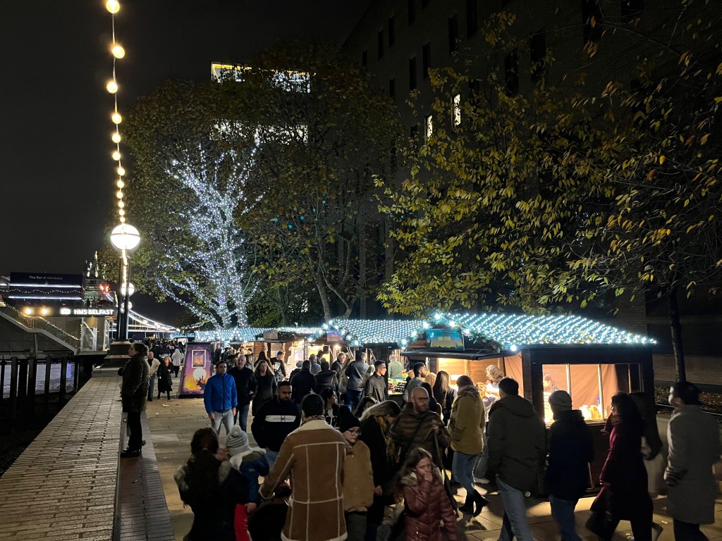 People shopping at street food stalls along the South Bank. The roofs of the stalls, along with a few tall trees in the distance, are covered in bright white lights to make them look festive.