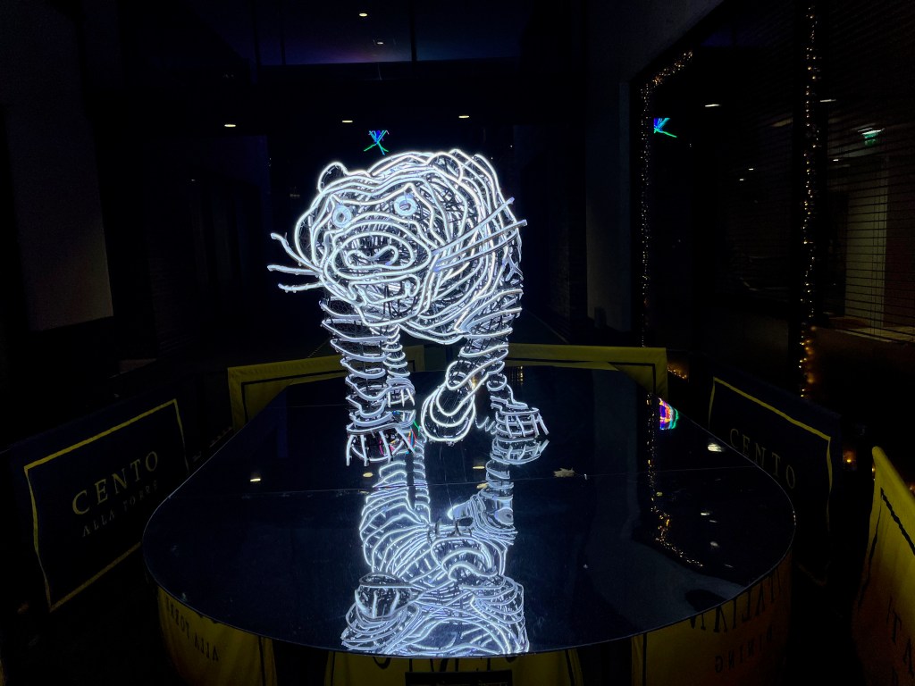 Head-on view of a sculpture of a tiger with long whiskers, made up of bright white, long curving string lights, standing on a mirrored base.