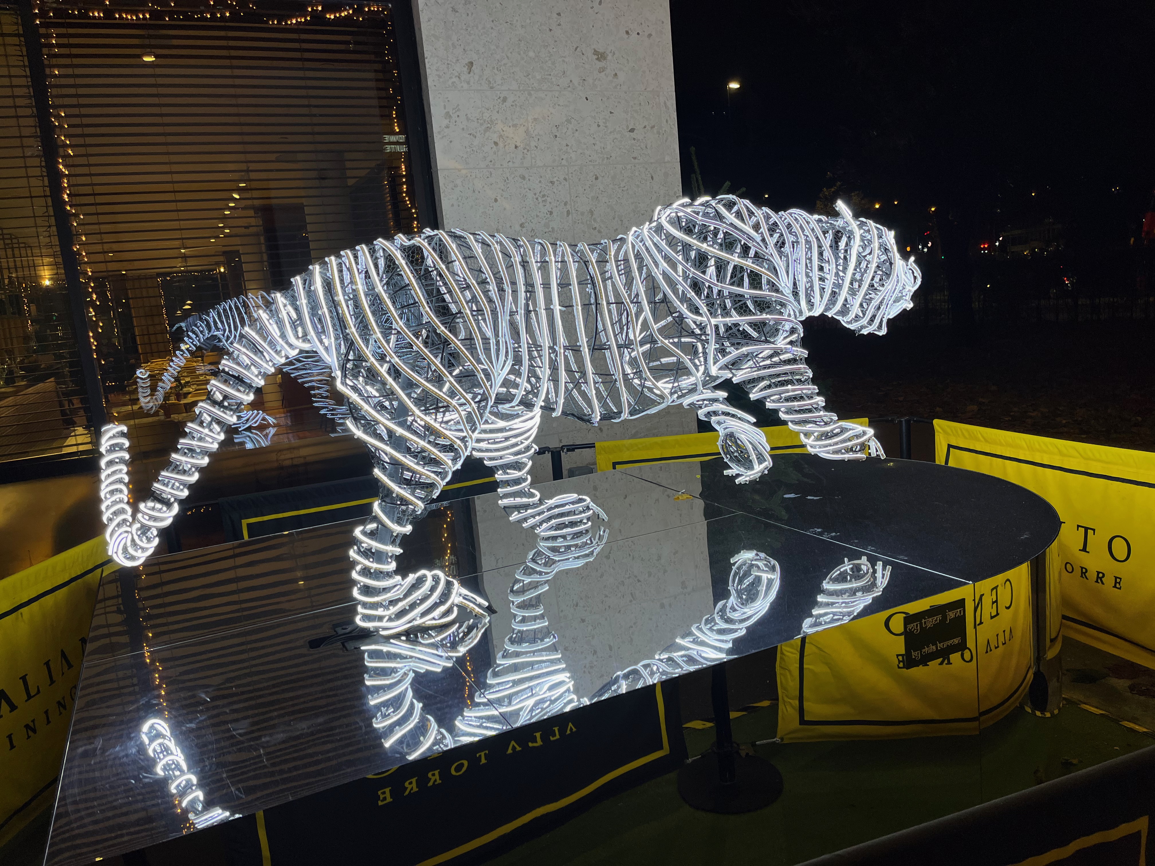 Rear view of a sculpture of a tiger with a long tail, made up of bright white, long curving string lights, standing on a mirrored base.