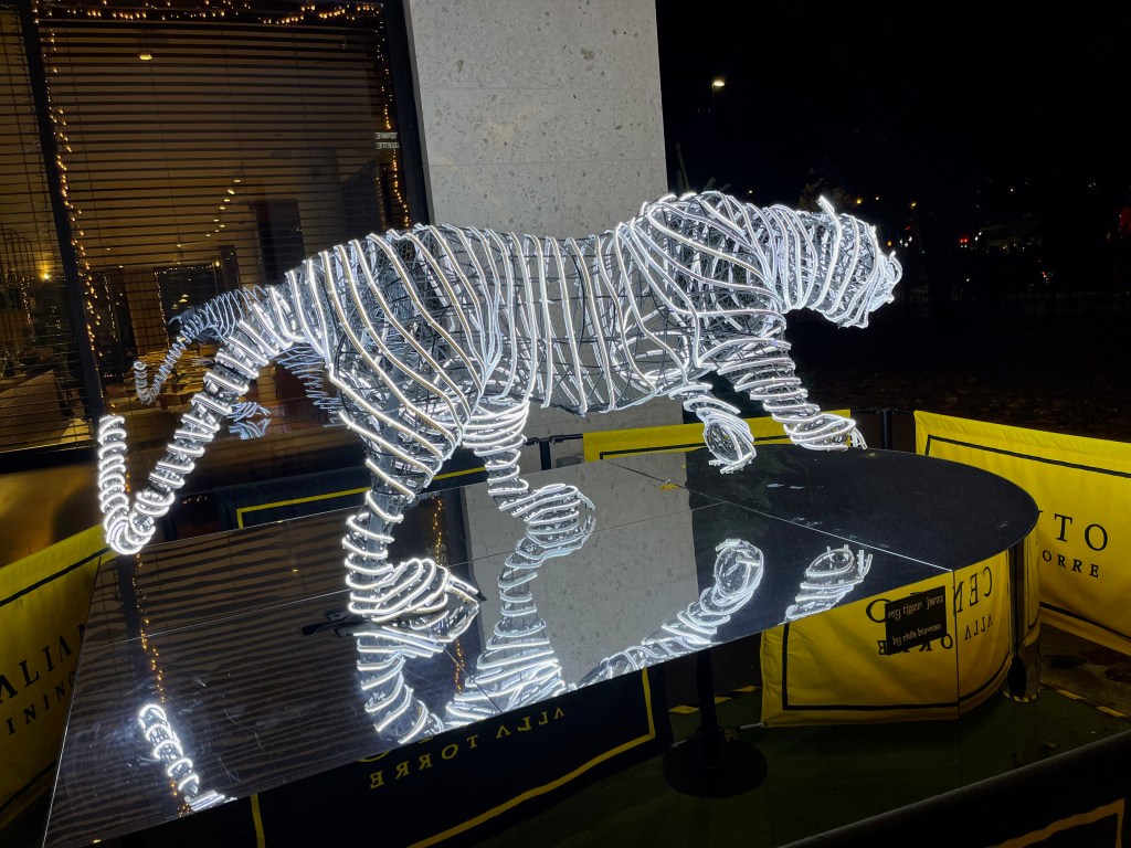 Rear view of a sculpture of a tiger with a long tail, made up of bright white, long curving string lights, standing on a mirrored base.
